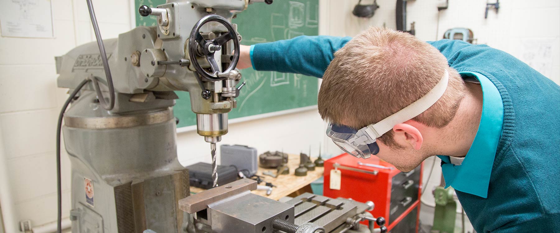 A physics student, wearing lab goggles, watches closely as he uses a drill press in a physics lab.
