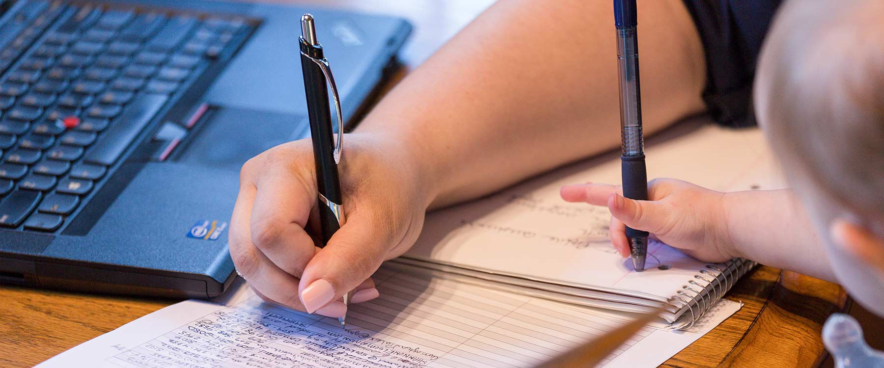 A continuing studies student writes information into a table. A child's hand holds a pen and scribbles in the student’s notebook.