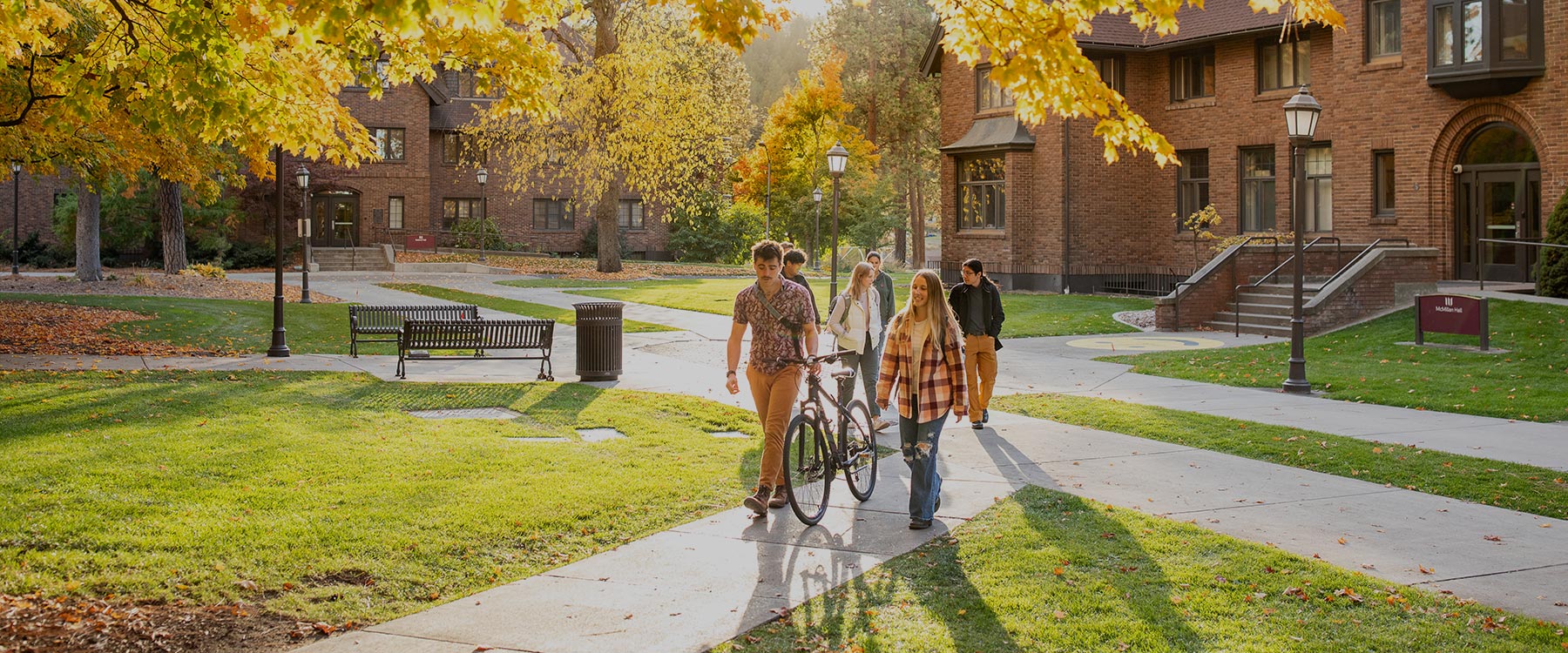 Many students walk between academic buildings on campus pathways beneath trees with fall foliage.