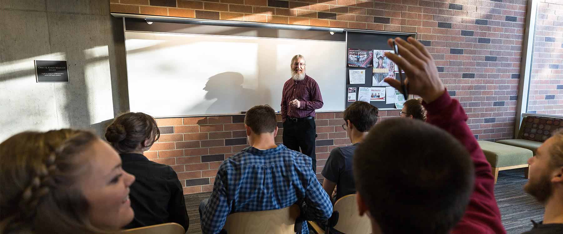 A professor stands in front of a whiteboard in a classroom, engaging with students, one of whom is raising their hand.