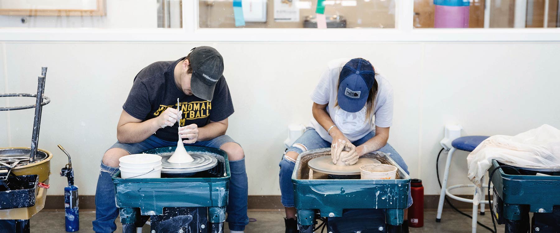 Two students seated at pottery wheels, shaping clay in a workshop. Both wear casual clothing and baseball caps.
