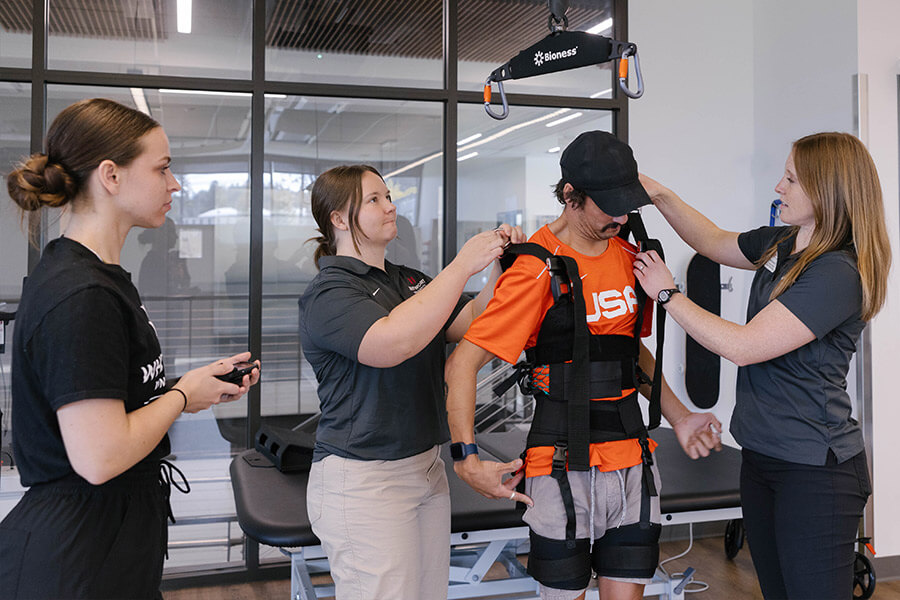 Two students adjust harness straps on a patient who is standing while another student observes.