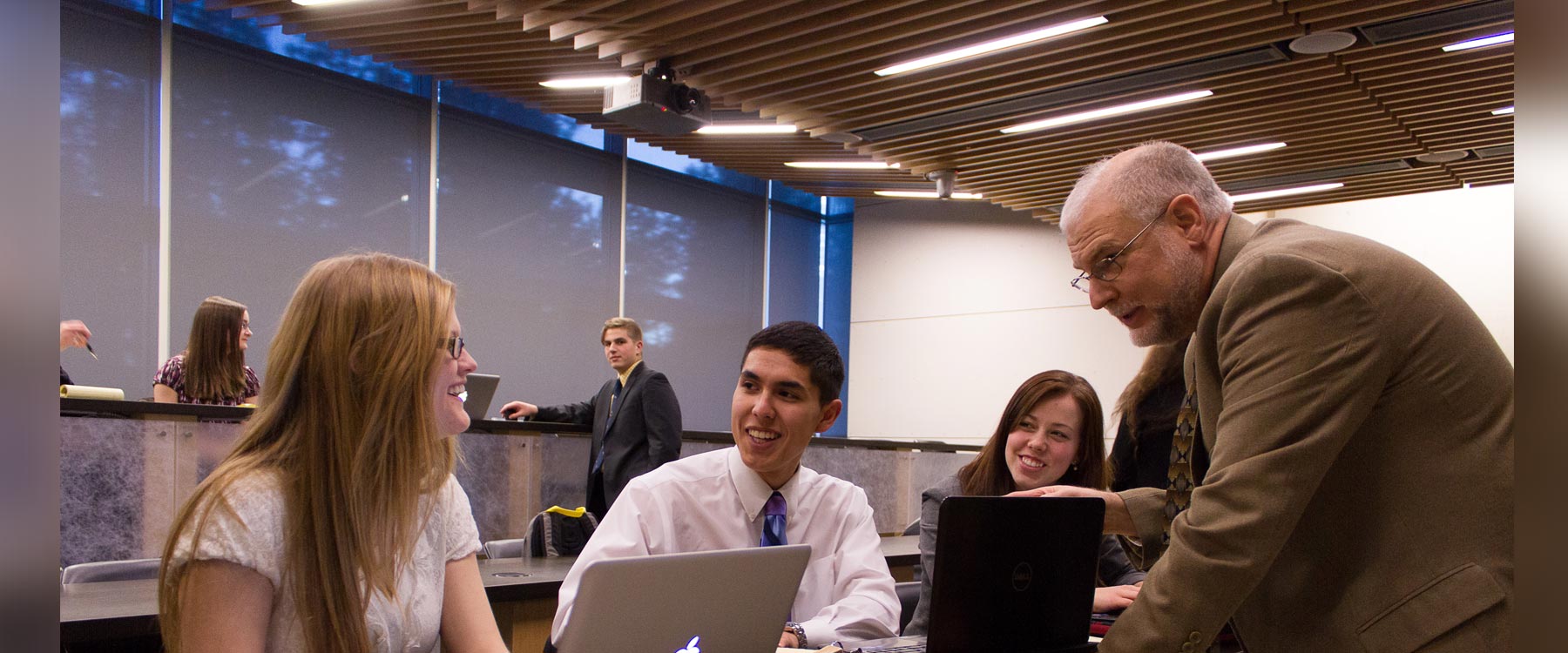 A professor converses with three students working on laptops in a modern classroom.