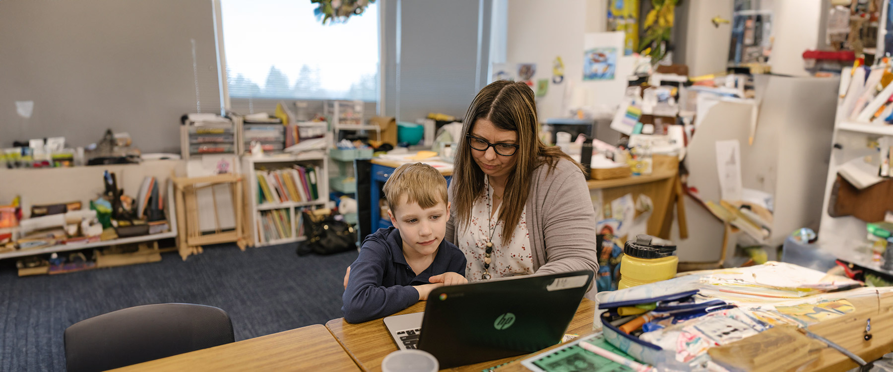 A teacher helps an elementary student use a laptop in a colorful classroom.