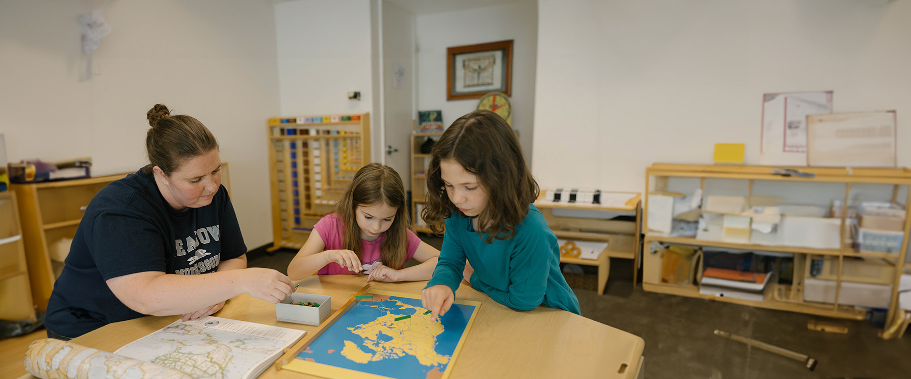 A teacher and two elementary students, seated at a table, work on a world map puzzle in a classroom.