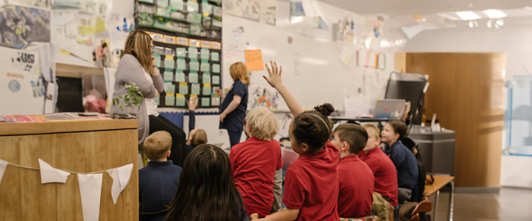 An elementary student raises their hand while another writes on a whiteboard observed by a teacher and peers.