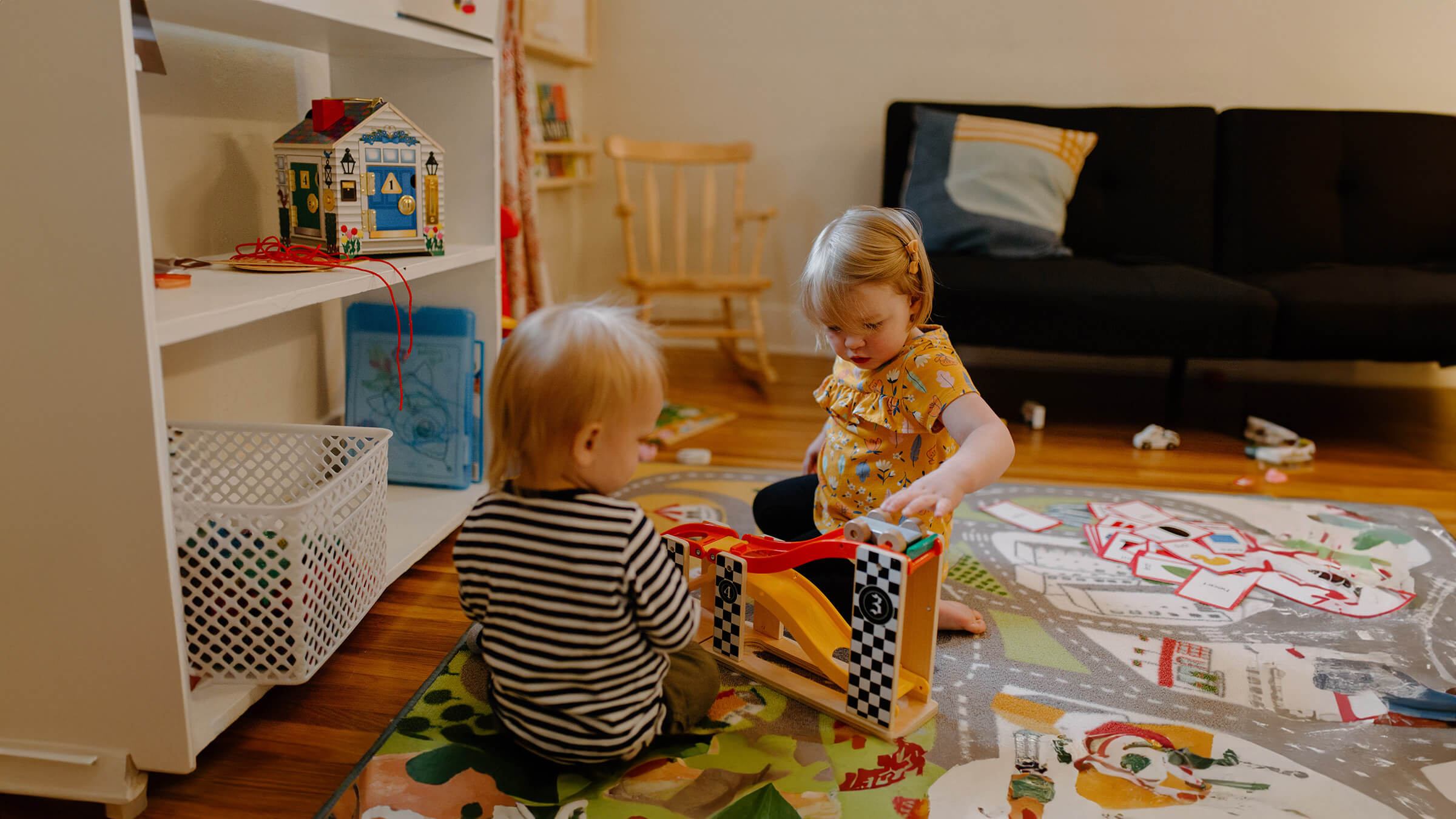 Two toddlers play with toy cars on a colorful mat in a classroom setting.