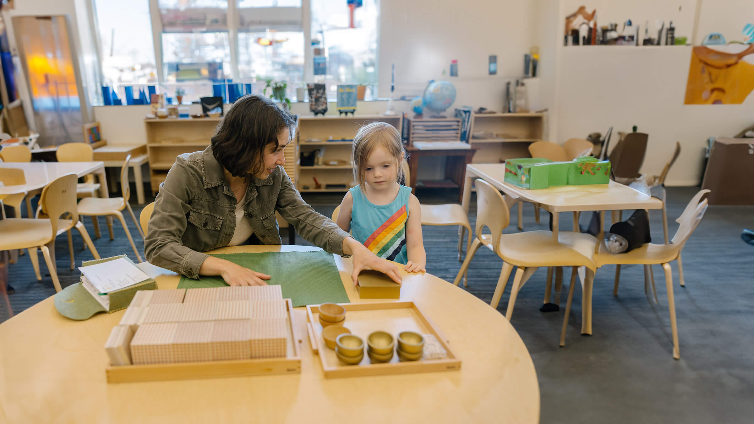 A teacher and young student work together at a table in a classroom with educational materials and art supplies.