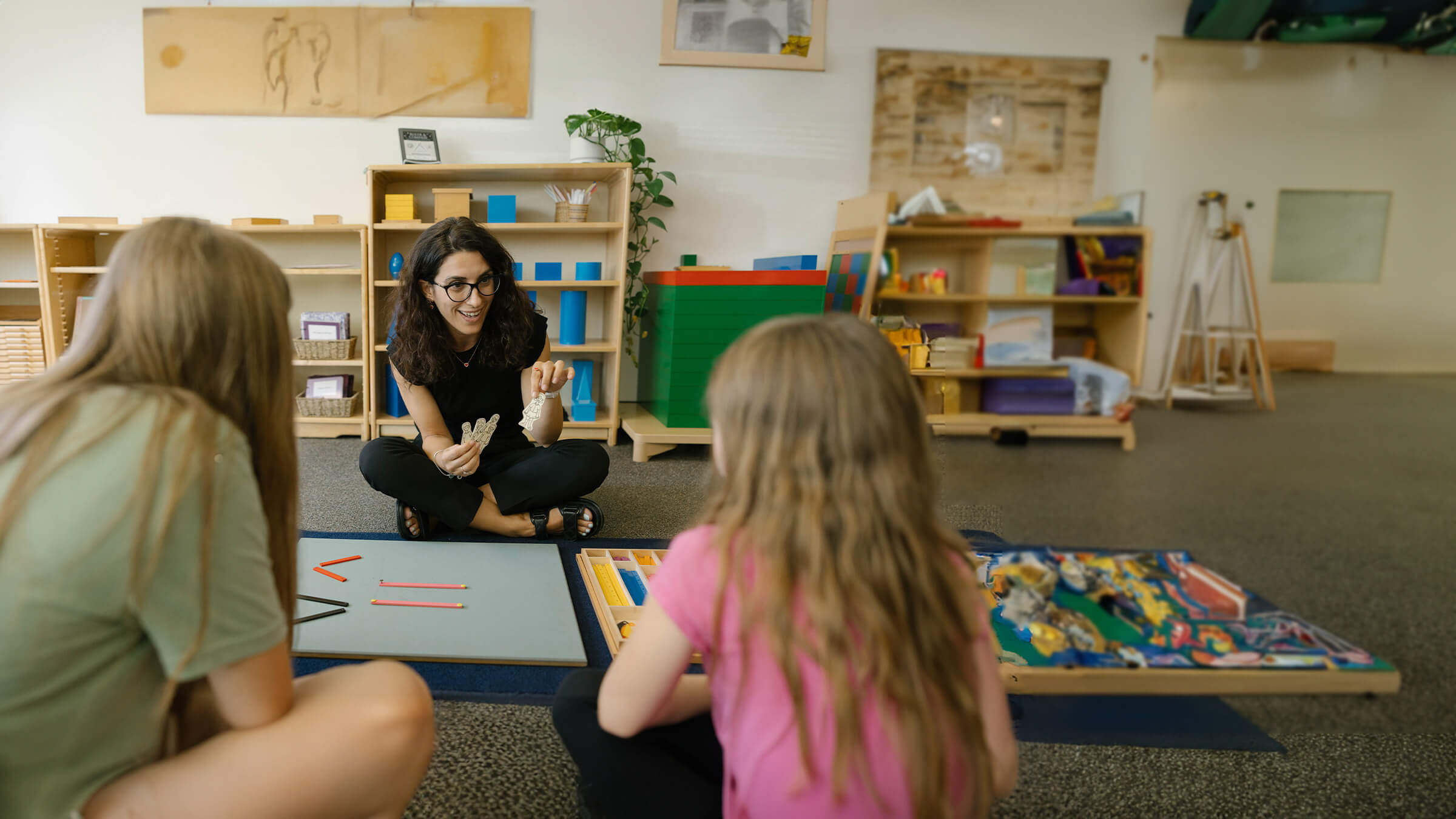 A teacher sits across from two elementary students on the floor and engages them with cutout objects.