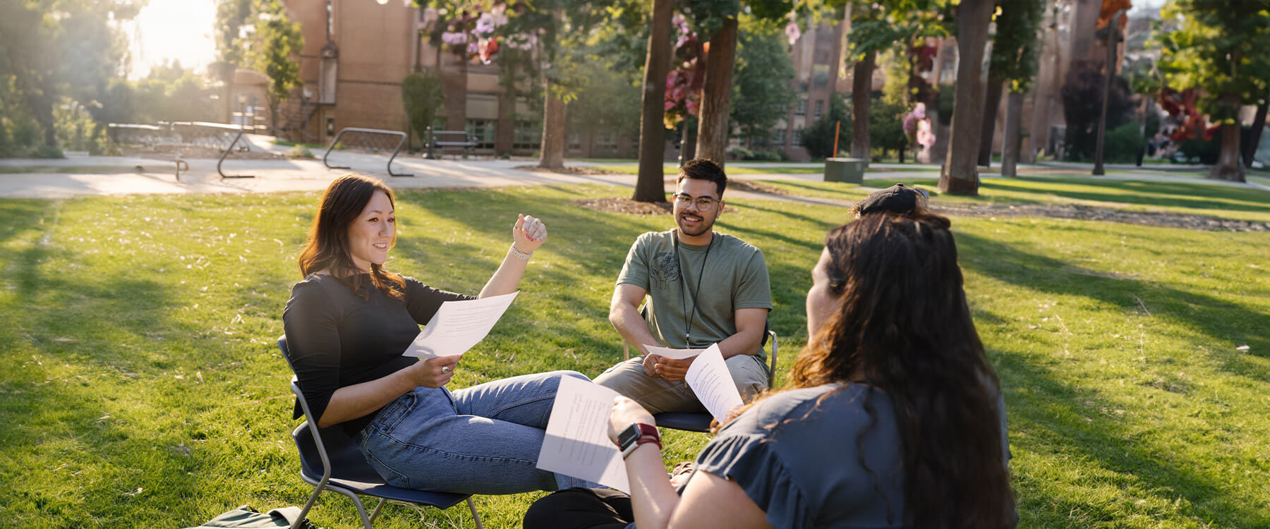 Four students sit outdoors in camp chairs, smiling and holding papers, with trees and buildings in the background.