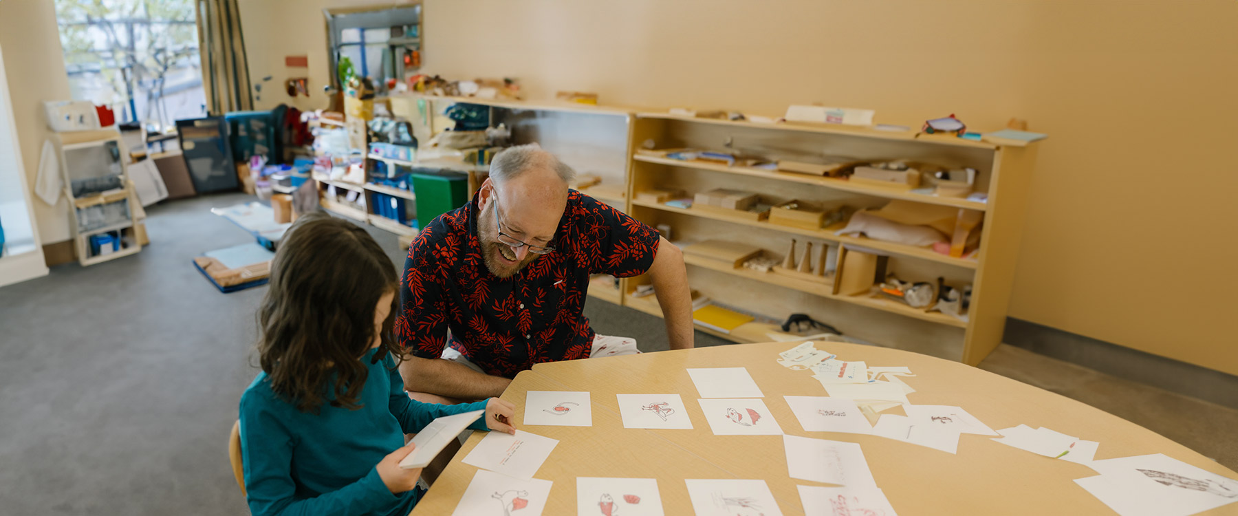 A teacher and a child sit at a table, engaging with drawings and cards, in an elementary classroom.