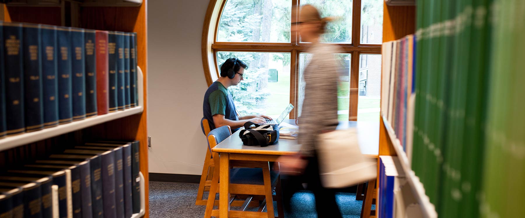 Person studying at a library table near a round window; another person walking past the bookshelves.