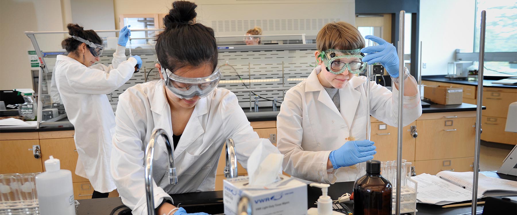 Students in a lab wearing white coats and goggles conducting experiments with various lab equipment.