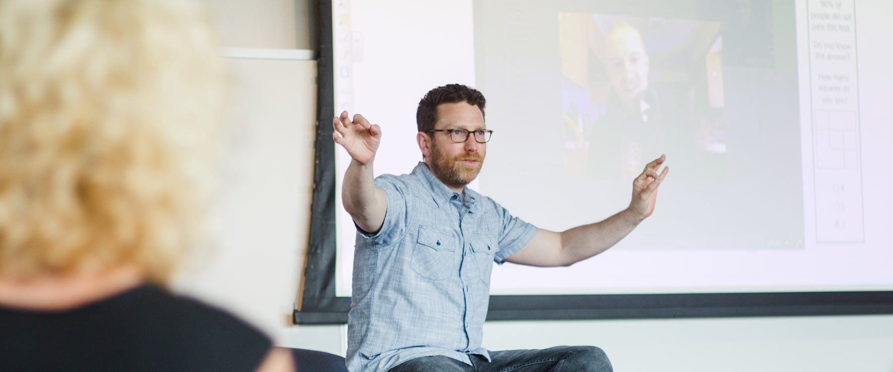 A professor gestures with both arms in front of a projector screen, presenting a lecture.