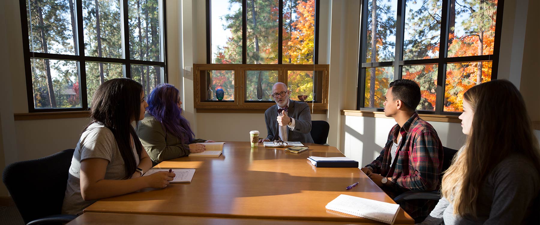 A professor sits at the end of a long table with students on either side listening intently.