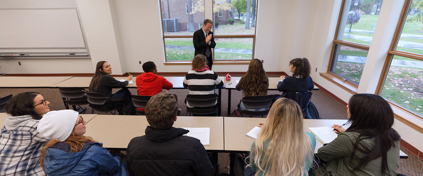 A professor laughs and claps his hands while addressing a group of students in a classroom.