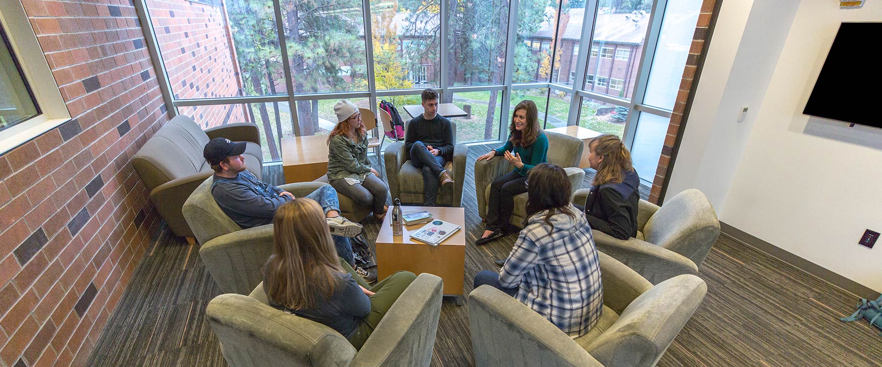 A professor sits with a group of students in a circle of armchairs near tall windows overlooking campus.
