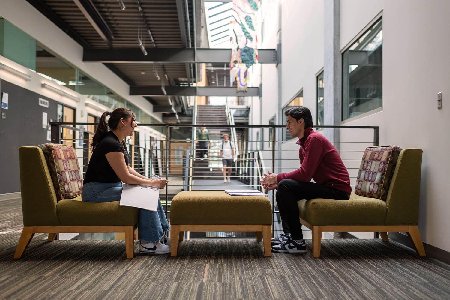 Two people engage in conversation while seated across from each other in a lounge area, set in an open hallway.