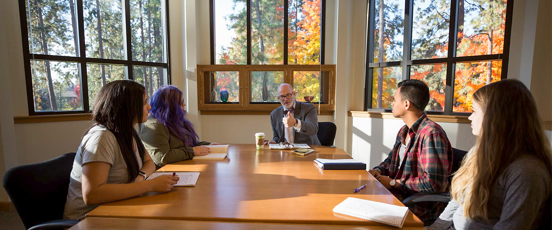 A professor sits at the end of a long table with students on either side listening intently.