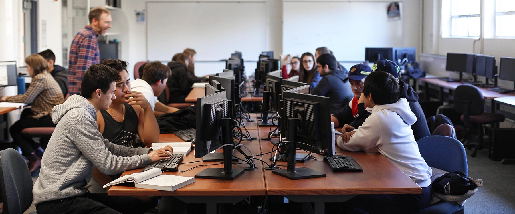 An instructor walks between rows of students in a computer lab.