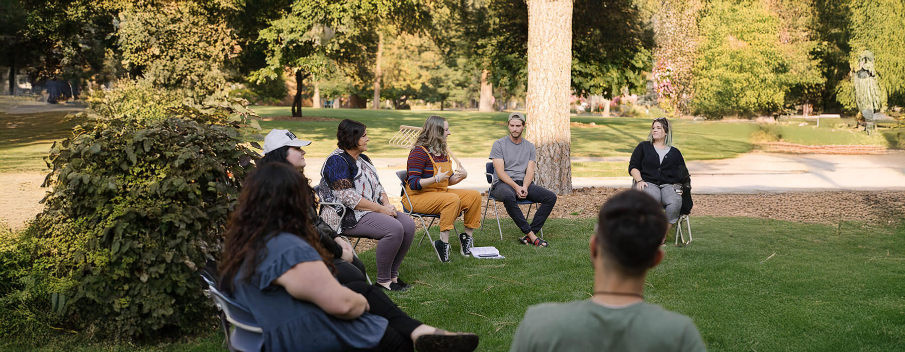 A group of adults sits outdoors in the shade of pine trees, engaging in a lively discussion.