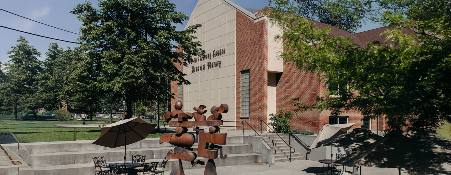 Building with brick and beige walls, surrounded by trees and outdoor seating with umbrellas, under a clear sky.