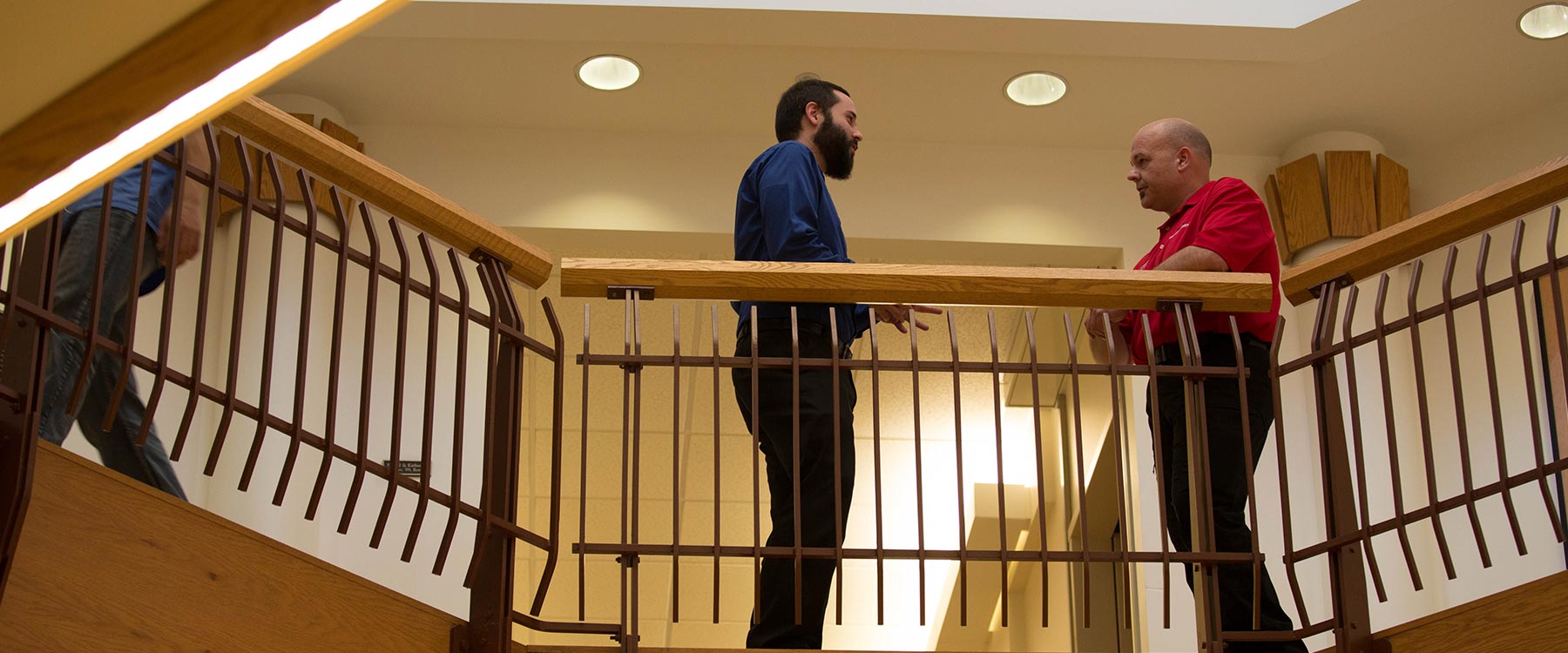 A student stands talking with a professor near a balcony railing in a warmly lit academic building.