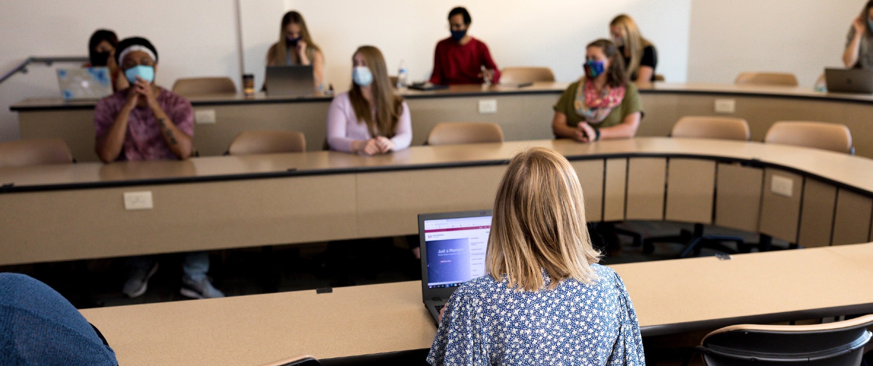 Students wearing masks and sitting in a classroom, with one person at a laptop facing them, leading the discussion.