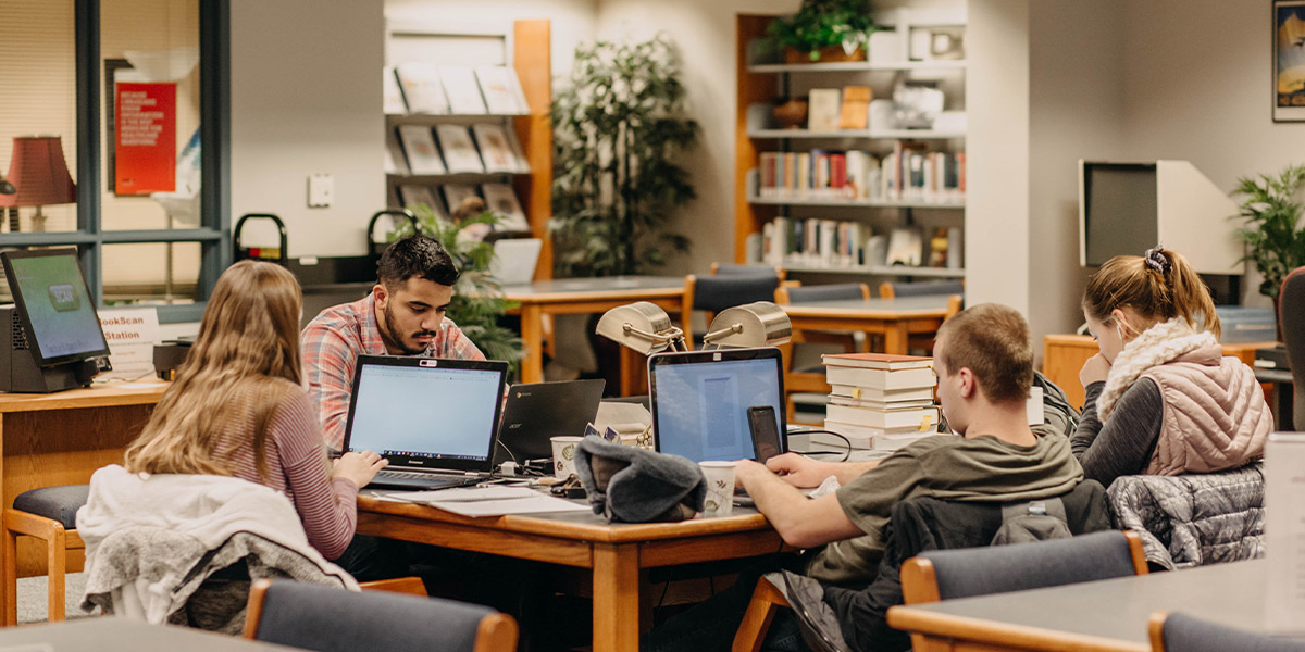Four students share a table in a library, three studying on laptops, one studying with a stack of books.