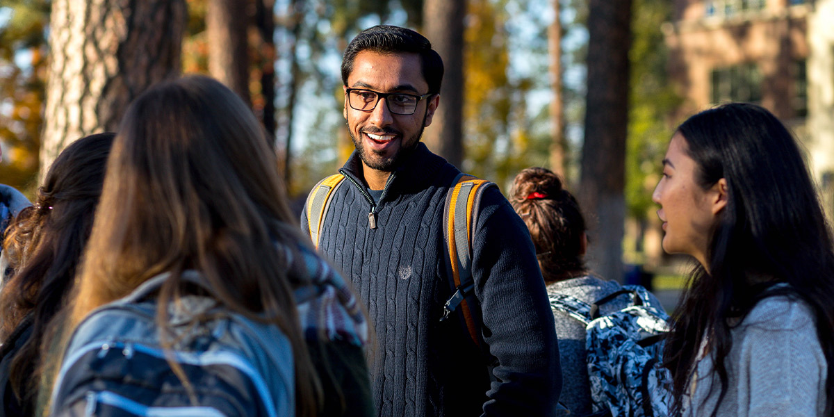 Three students wearing fall clothing and backpacks chat with each other outdoors in a crowd on a sunny day.