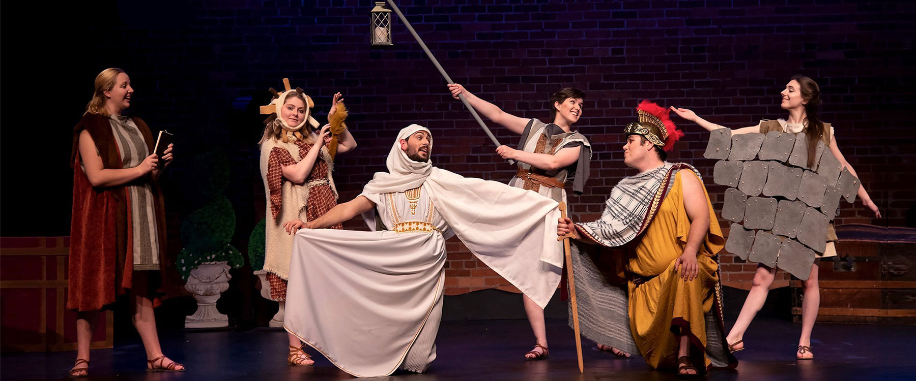 A lively theater scene with actors in medieval costumes performing on stage against a brick backdrop.
