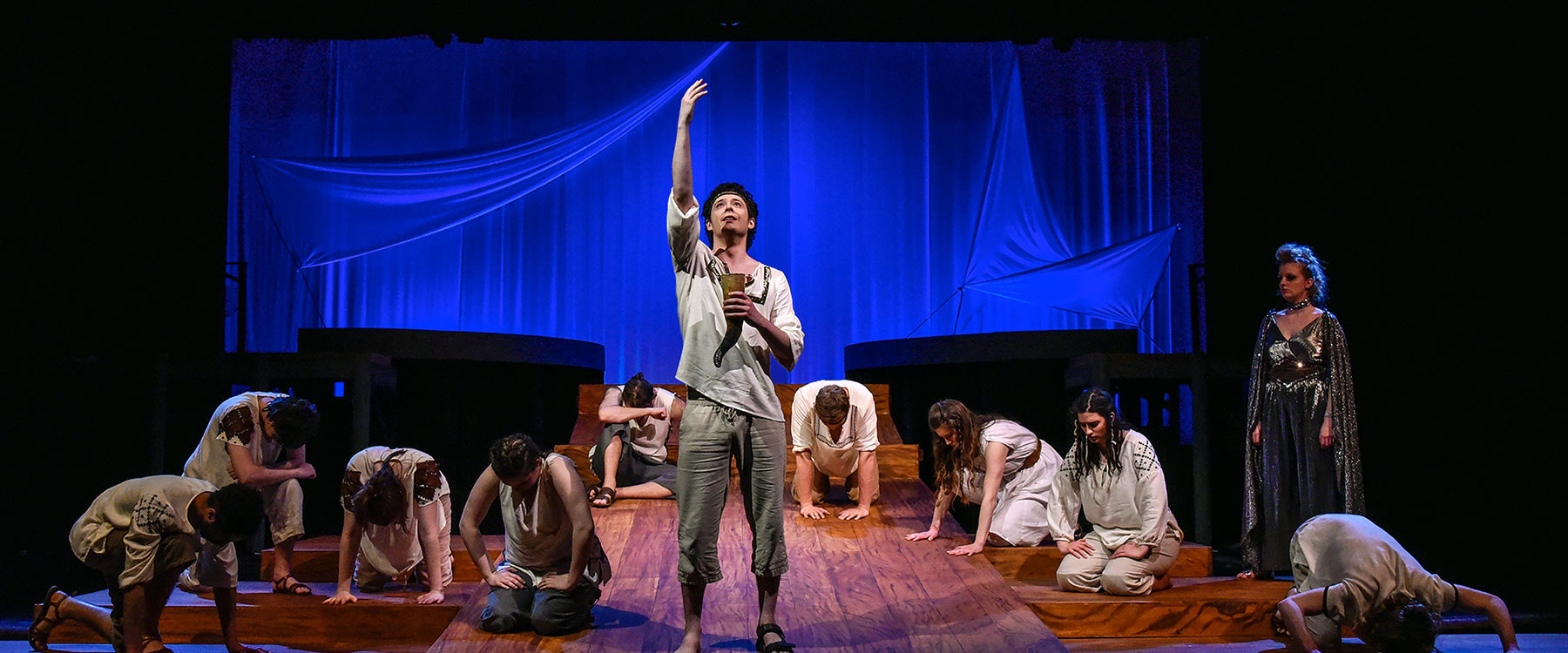 Actors perform a theatrical scene on stage; one stands and reaches upward, while others kneel.
