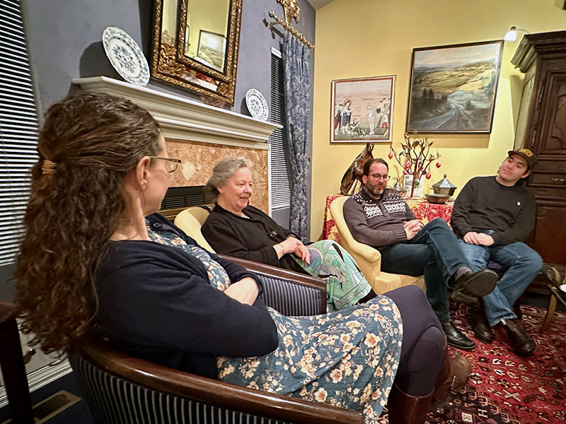 A few students and professors gather in comfortable chairs around a cozy living room.