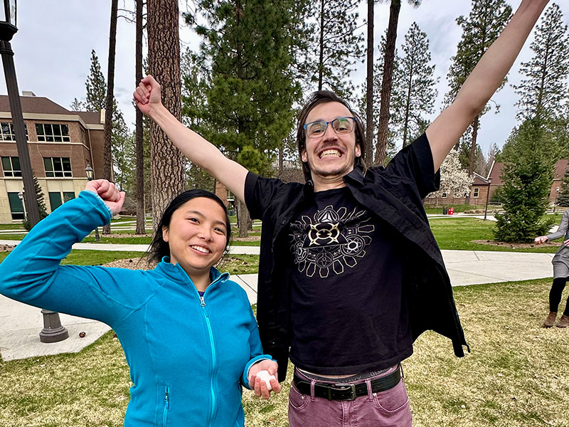 Two students stand outdoors, smiling and celebrating with raised arms. One holds an egg.