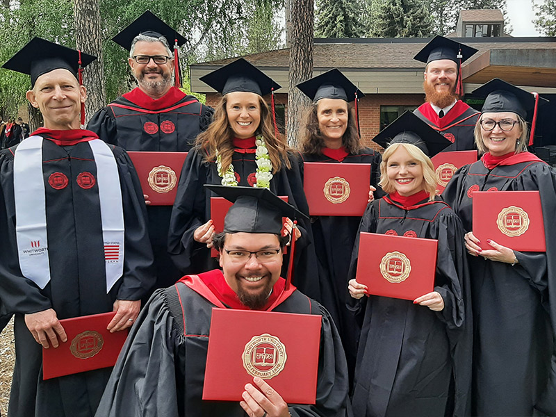 A group of eight graduates in caps and gowns smiles while holding their diplomas.