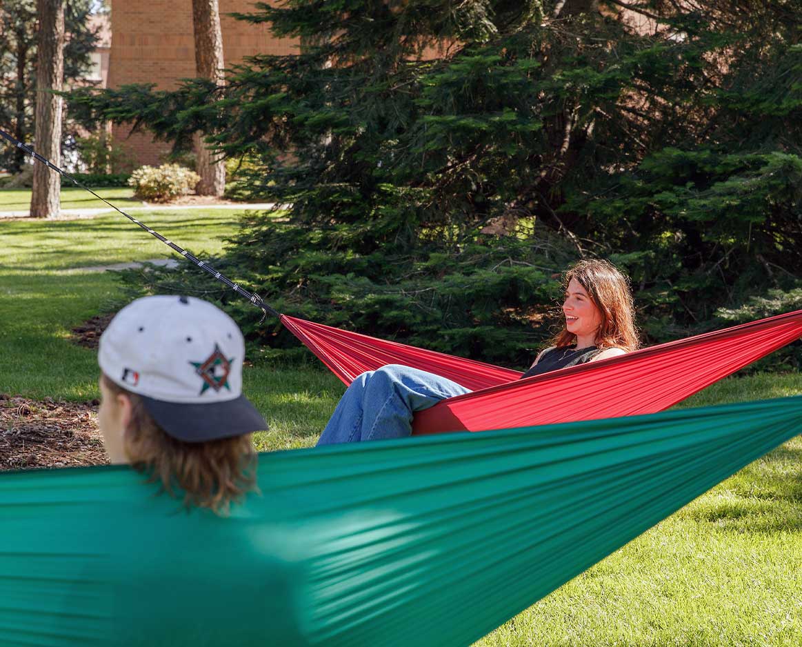 Two students relax in hammocks hung between pine trees on campus.