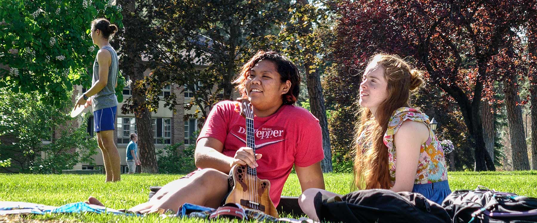 Two students sit in the grass and two are seen in the distance, playing Frisbee, on a warm day.