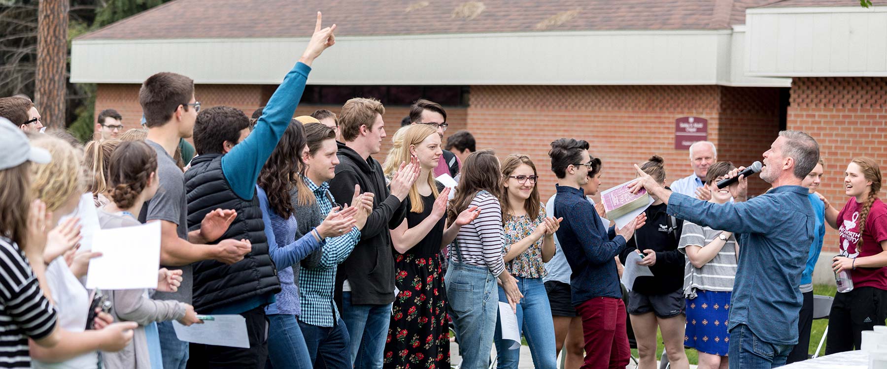 Students smile and clap as a professor addresses them through a microphone at an outdoor gathering.