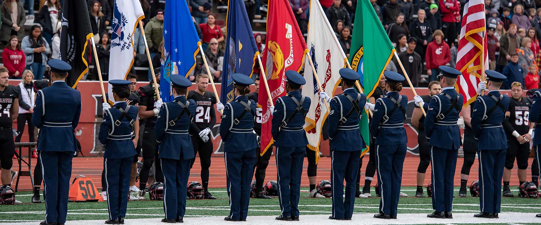 Uniformed officers hold flags, including the American flag, and football players stand in salute.
