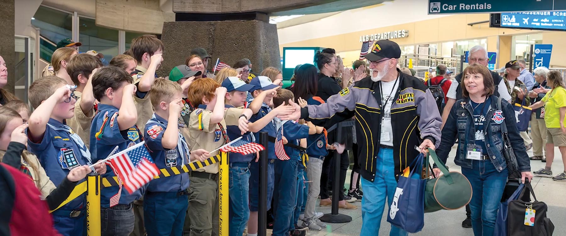 Boy Scouts enthusiastically welcome an elderly veteran at an airport, saluting and giving high-fives.