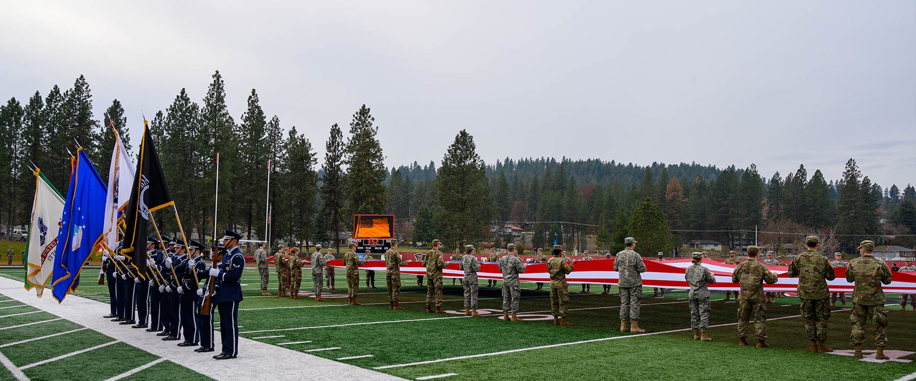 Officers stand with flags near a football field as military members hold a large American flag across the field.