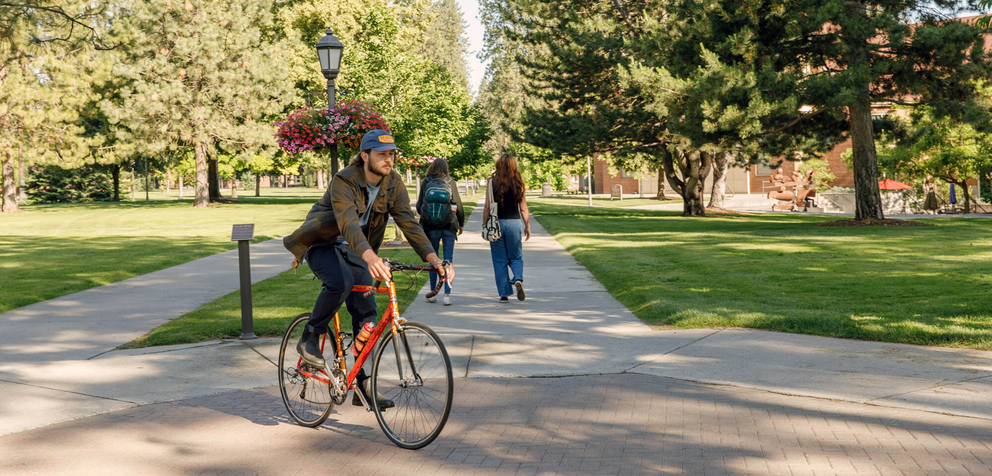 A student rides a red bicycle on a paved path through manicured lawns and evergreen trees past a brick academic building.