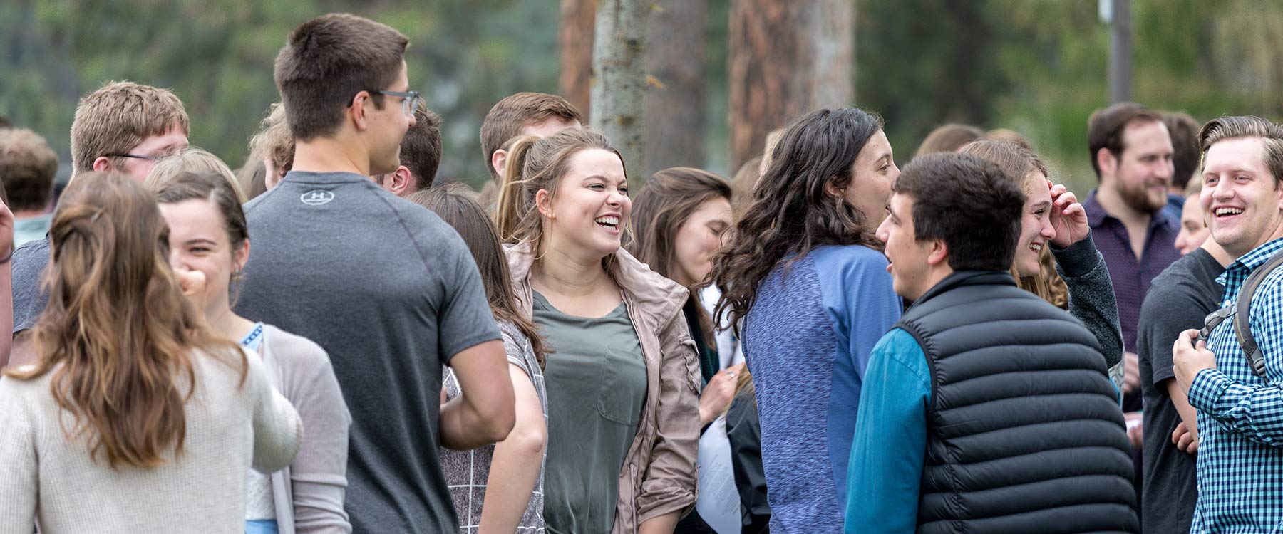 A large group of students smiling and conversing outdoors at a campus event beneath pine trees.