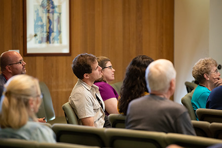 An attentive audience sits together, looking forward, in a room with wooden walls.