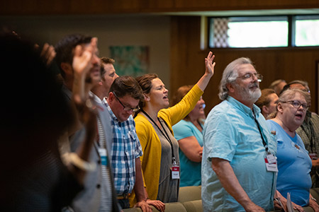 Several people stand in a group, some with hands raised, participating in an indoor worship event.