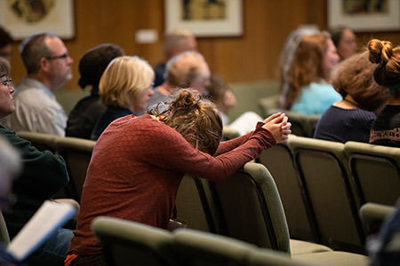 A person in a red sweater sits, leaning forward in prayer, surrounded by others in a seated gathering.