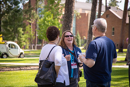 Three people, one wearing a lanyard, chat and laugh together outdoors on a sunny day, with trees and brick buildings in the background.