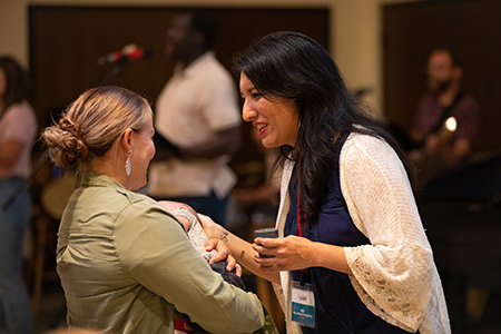 Two people, one holding a baby, smile and talk with one another in a large room with a musician in the background.