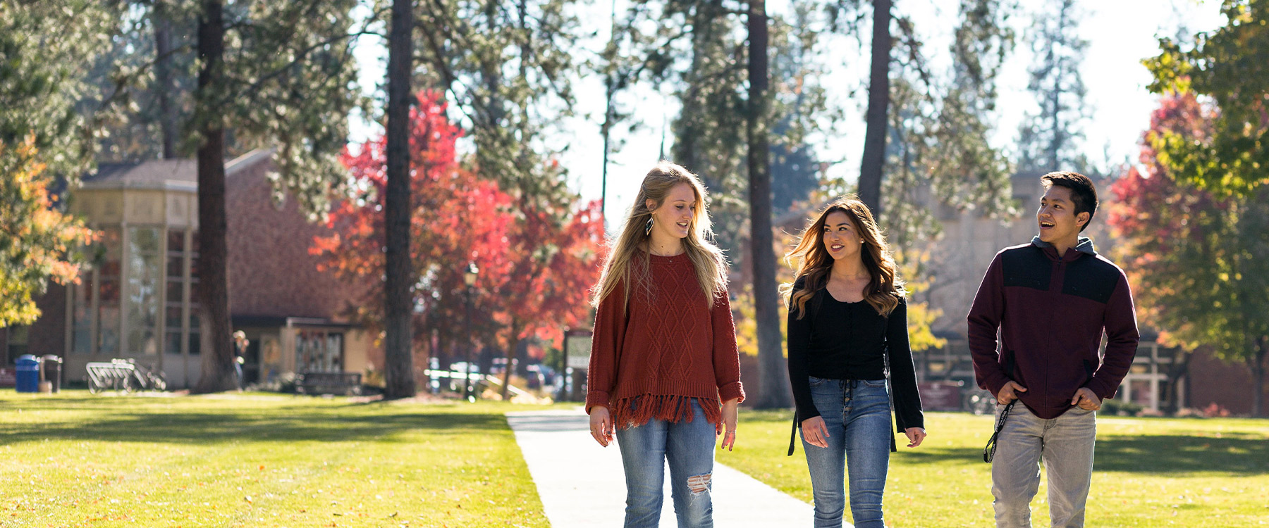 Three students walk outdoors on a tree-lined path with fall foliage, engaged in conversation and smiling.