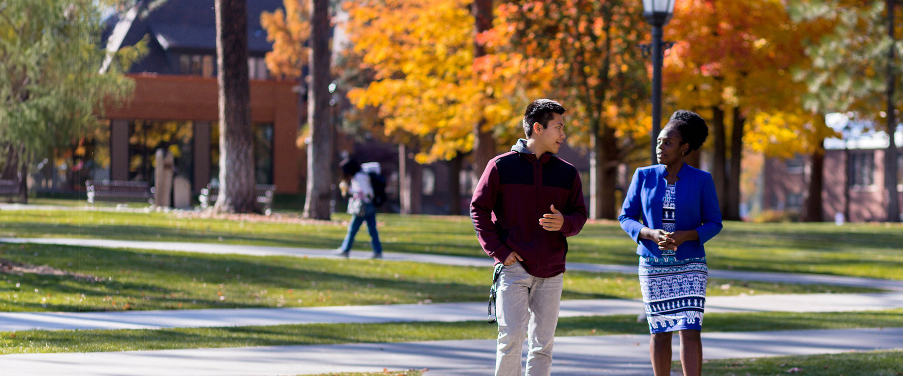 A professor and student walk down a pathway lined with trees in autumn with academic buildings in the background.